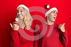 Young interracial couple wearing christmas hat smiling with happy face looking and pointing to the side with thumb up