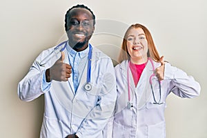 Young interracial couple wearing doctor uniform and stethoscope doing happy thumbs up gesture with hand