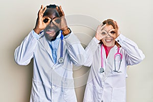 Young interracial couple wearing doctor uniform and stethoscope doing ok gesture like binoculars sticking tongue out, eyes looking