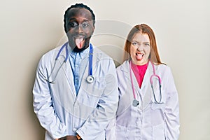 Young interracial couple wearing doctor uniform and stethoscope sticking tongue out happy with funny expression