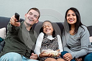 Young interracial family gawking at the TV from the couch. Young caucasian father control remote and daughter hold popcorn