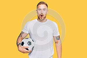 Young irish man holding soccer ball scared and amazed with open mouth for surprise, disbelief face