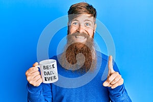 Young irish redhead man drinking mug of coffe with best dad ever message smiling with an idea or question pointing finger with