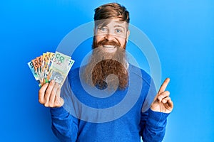 Young irish redhead man holding australian dollars smiling happy pointing with hand and finger to the side