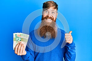 Young irish redhead man holding hong kong 50 dollars banknotes pointing thumb up to the side smiling happy with open mouth