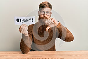 Young irish redhead man holding paper with insult message sititng on the table with angry face, negative sign showing dislike with