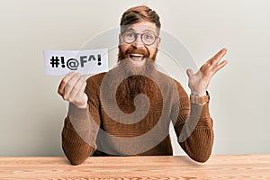 Young irish redhead man holding paper with insult message sititng on the table celebrating victory with happy smile and winner