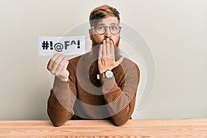 Young irish redhead man holding paper with insult message sititng on the table covering mouth with hand, shocked and afraid for