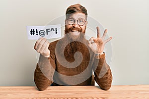 Young irish redhead man holding paper with insult message sititng on the table doing ok sign with fingers, smiling friendly