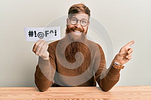 Young irish redhead man holding paper with insult message sititng on the table smiling happy pointing with hand and finger to the