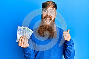 Young irish redhead man holding south african 100 rand banknotes pointing thumb up to the side smiling happy with open mouth