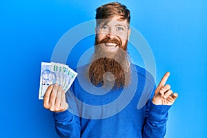 Young irish redhead man holding south african 100 rand banknotes smiling happy pointing with hand and finger to the side