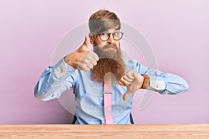 Young irish redhead man wearing business shirt and tie sitting on the table doing thumbs up and down, disagreement and agreement