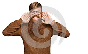 Young irish redhead man wearing casual clothes and glasses trying to hear both hands on ear gesture, curious for gossip