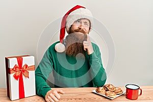 Young irish redhead man wearing christmas sitting on the table with gift serious face thinking about question with hand on chin,