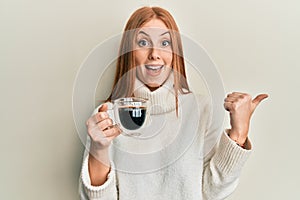 Young irish woman drinking a cup of coffee pointing thumb up to the side smiling happy with open mouth