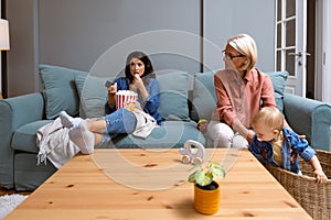 Young irresponsible mother watch TV and eat popcorn while her mother child grandmother looks after the baby. Carefree mom pays no