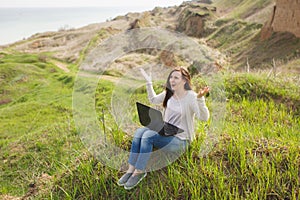 Young irritated dissatisfied business woman or student in casual clothes sitting on grass using laptop in field