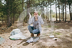 Young irritated woman in casual clothes and gloves cleaning rubbish and spreading hands near trash bags in park.Problem