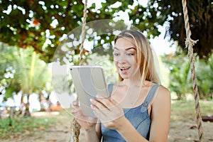 Young jocund woman using tablet and riding swing on sand, wearing jeans sundress.