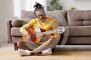 Young joyful african american man playing acoustic guitar at home, sitting on floor in living room