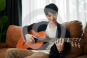 Young joyful Asian man playing guitar while sitting in the sofa in his free time