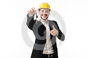 Young joyful businessman in yellow safety helmet showing thumb up happily looking in camera with keys of new flat in