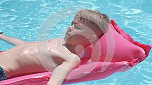 Young joyful child boy having fun swimming on inflatable air mattress in swimming pool with blue water on warm summer