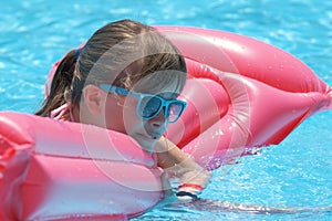 Young joyful child girl having fun swimming on inflatable air mattress in swimming pool with blue water on warm summer
