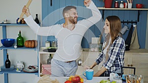 Young joyful couple have fun dancing and singing while set the table for breakfast in the kitchen at home