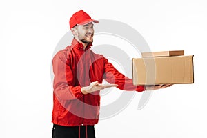 Young joyful delivery man in red cap and jacket happily giving package to customer over white background isolated