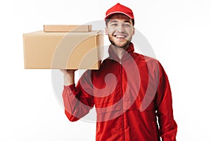 Young joyful delivery man in red cap and jacket holding parcel in hand happily looking in camera over white background