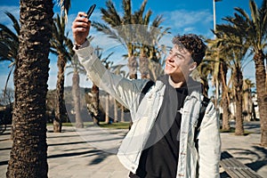 Young joyful man happily taking selfie on smartphone during walk through park with palm trees