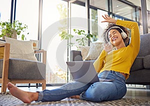 Young joyful mixed race woman wearing headphones and listening to music while using a laptop at home. One cheerful