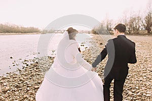 Young just married couple walking back holding hands on the river coast