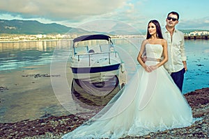 Young just married couple in wedding gown and suit standing near the boat at the seaside looking aside