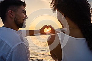 Young just married multiracial couple in love making heart shape with hands with setting sun in centre of it, looking in the eyes