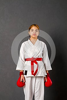 Young karateka girl in a white kimono and red competition outfit trains and performs a set of exercises against a gray wall