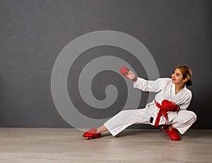 Young karateka girl in a white kimono and red competition outfit trains and performs a set of exercises against a gray wall