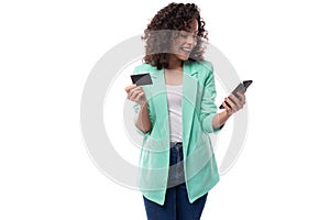young kind curly brunette assistant woman dressed in an office blue jacket uses a smartphone and a bank card to transfer