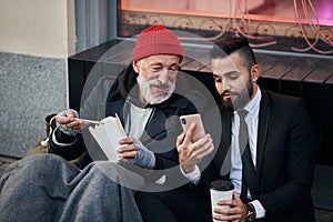 Young kind-hearted businessman and senior beggar look at phone