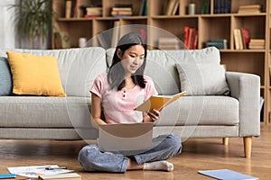 Young korean lady studying online from home, reading book and using laptop pc, sitting on floor near sofa, free space