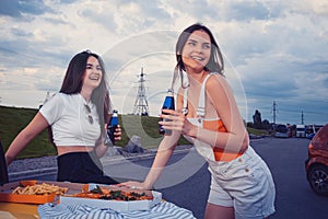 Young ladies are smiling, enjoying soda in glass bottles, posing leaning on trunk of yellow car with pizza and french