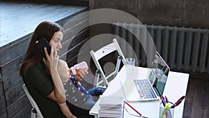 Young lady is calling by phone, sitting on a chair and holding baby on a knees.