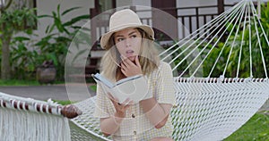 Young lady in hat with book in hammock. Charming happy woman resting in hammock and reading book at green garden. Fair