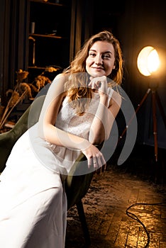 A young lady, her long hair cascading, sits gracefully in a chair, smiling warmly at the camera in the dim light of the