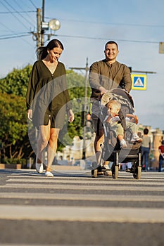 A young lady in khaki colour dress and her husband crossing over the road on zebra crossing with their baby boy sitting in a strol