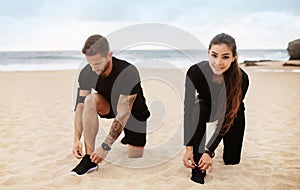 Young lady and man in sportswear tying shoelaces on shoes on ocean beach, getting ready for morning workout