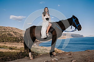 A young lady rider in a white dress is sitting in the saddle on a horse, against the background of a beautiful mountain