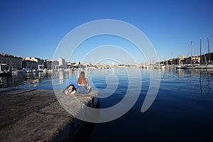 Young lady sitting alone by a harbor with her backpack next to her watching the ships and buildings